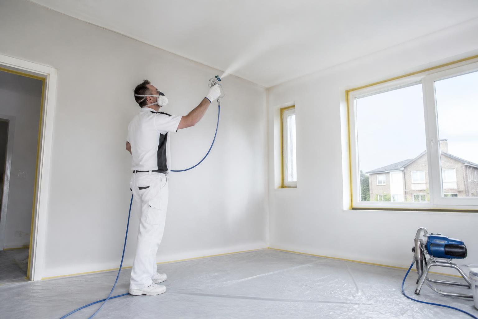 Vakman spuit wanden en plafond van een lege ruimte met airless apparatuur en afgeplakte vloer